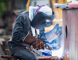 Photo shows welder wearing eye protection and gloves while welding a pipe connection.