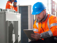 Photo shows worker with tablet computer making notes while inspecting an HVAC unit.